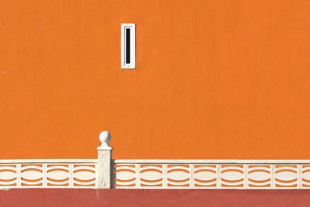 A simple orange wall with a small vertical window near the top and a white decorative fence at the bottom, creating a minimal composition.